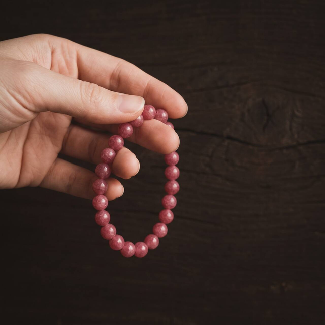 Deep rose-pink round-bead bracelet held in one hand against dark wood